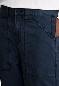 Pantaloni blu navy con una texture liscia, caratterizzati da tasche inclinate e cuciture visibili. Dettaglio con chiusura a bottone in vita.