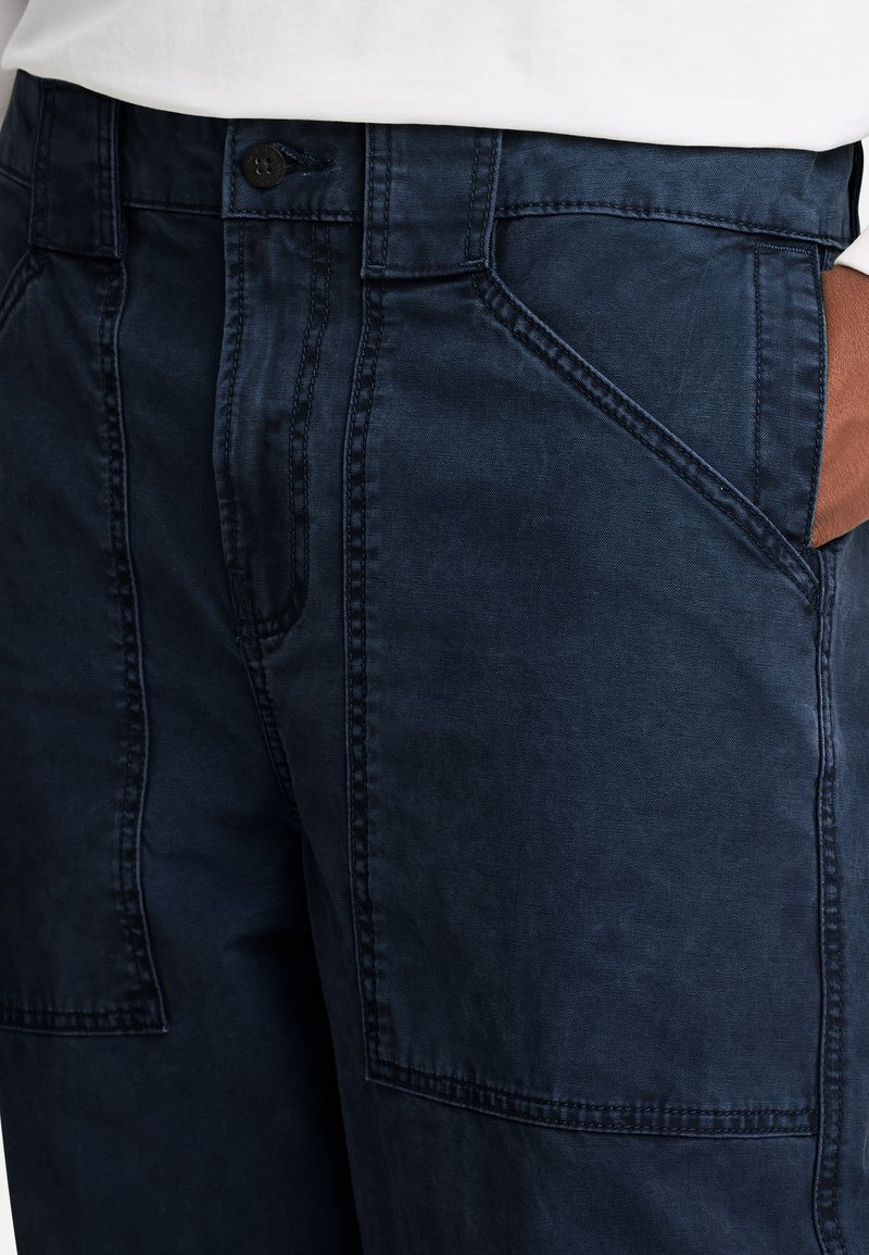 Marineblaue Hose mit glatter Textur, die über schräge Taschen und sichtbare Nähte verfügt. Knopfverschluss an der Taille.