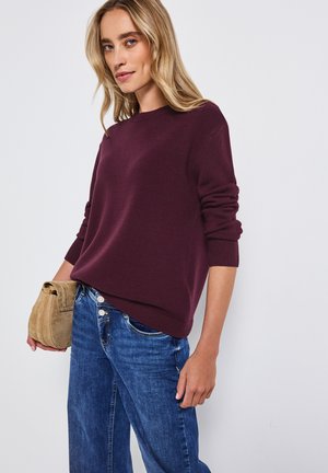 Pull en maille bordeaux à col rond et poignets côtelés, associé à un jean en denim bleu clair et un sac en cuir suédé beige porté en travers.