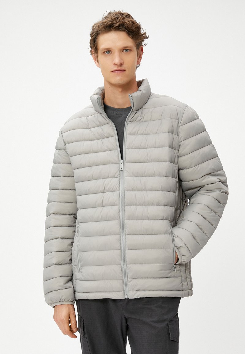 Chaqueta de plumas gris claro con un cuello alto, acolchada horizontalmente, cremallera frontal y bolsillos laterales, hecha de un tejido suave y resistente al agua.