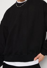 Černá mikina s texturovanou látkou, volným střihem a žebrovanými manžetami a spodním lemem. V kombinaci s vrstvenou bílou košilí pod ní.