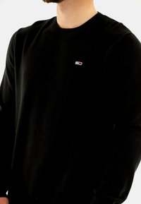 Svart stickad tröja med rund halsringning och ribbade ärmslut. Har en liten logotyp i röd och vit på vänster bröst.