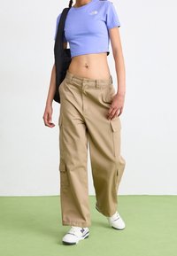Pantalons cargo beige avec de multiples poches, associés à un crop top violet clair et des baskets blanches. Le haut présente un petit logo.