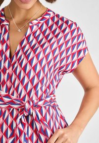 Värikäs mekko, jossa on geometrinen timanttikuvio punaisena, sinisenä ja valkoisena. Lyhyet hihat, napitus etuosassa, vyötärösolki. Silkkinen kankaan rakenne.