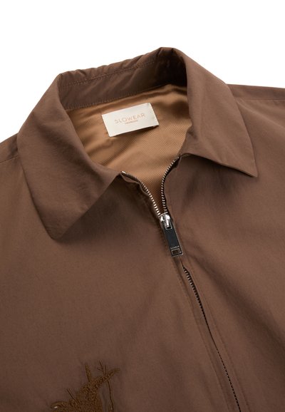 Giacca marrone con colletto, cerniera argento e ricamo vicino al fondo, etichetta del marchio "SLOWEAR" visibile all'interno del collo.