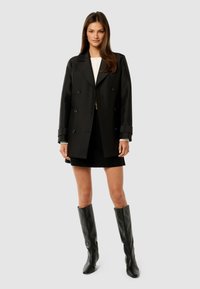 Young woman wearing a black double-breasted coat, black skirt, white top, and black knee-high boots, standing against a plain light background.