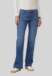 Jean en denim bleu clair avec une coupe évasée, cinq poches et des détails surpiqués. Porté avec un haut blanc et des baskets crème.