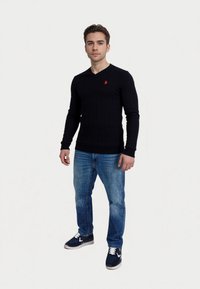 Tengerészeti bordázott kötött V nyakú pulóver hosszú ujjakkal, kis piros logóval; világoskék farmernadrággal és tengerészkék sportcipővel párosítva.