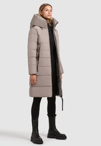 Bežový prešívaný kabát s vysokým golierom a bočnými zipsovými vreckami. Nosený cez čierne legíny a masívne čierne čižmy. Hladká textúra.