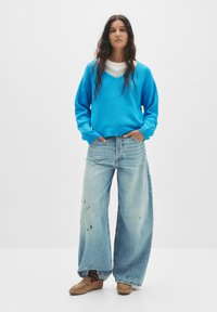 Hellblauer, gestrickter Pullover mit V-Ausschnitt, getragen über einem grauen, geschichteten Hemd; kombiniert mit übergroßen, lädierten hellen Jeans und braunen Mokassins.
