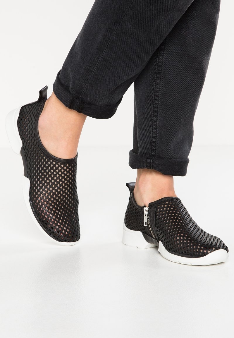 Personne portant des chaussures noires sans lacets avec un design perforé respirant et des semelles blanches, associées à un pantalon noir retroussé.