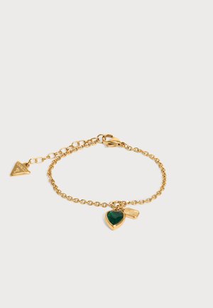 Bracelet chaîne en or avec un pendentif en forme de cœur vert, un petit pendentif rectangulaire et un fermoir réglable avec une étiquette triangulaire au logo Guess.