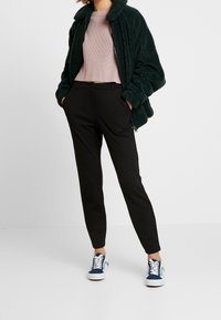 Groene fuzzy jas, roze geribde trui, zwarte tapered broek en blauwe sneakers met witte zolen. Opvallende textuur en kleurcontrast.