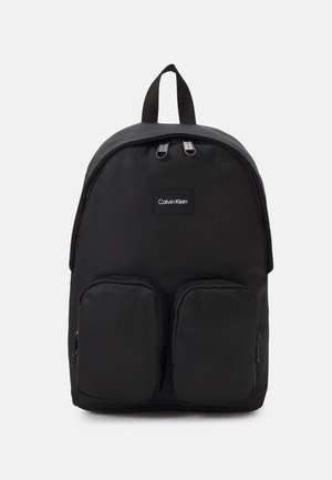 Zaino nero in nylon con due tasche frontali con zip, spallacci regolabili e maniglia superiore. Presenta una patch con il marchio al centro.