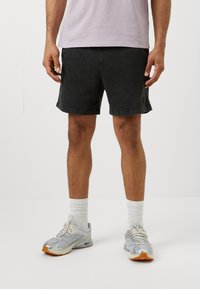 Black denim shorts with a relaxed fit, featuring a subtle faded texture and a side logo detail. Paired with light gray athletic shoes and white socks.