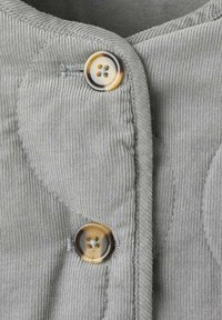 Il tessuto in corduroy grigio presenta prominenti coste orizzontali, con un bottone rotondo a quattro fori come dettaglio di chiusura. Le cuciture esaltano la texture.