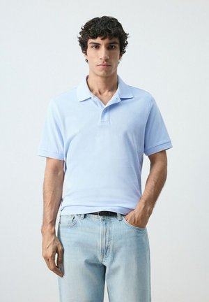 Giovane uomo con capelli ricci neri che indossa una polo azzurro chiaro e jeans di denim chiaro, in piedi davanti a uno sfondo semplice.