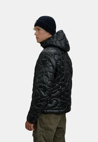 Giacca a vento nera con un design trapuntato e testurizzato e cappuccio. Presenta una toppa con logo laterale ed è abbinata a un berretto in maglia scuro.