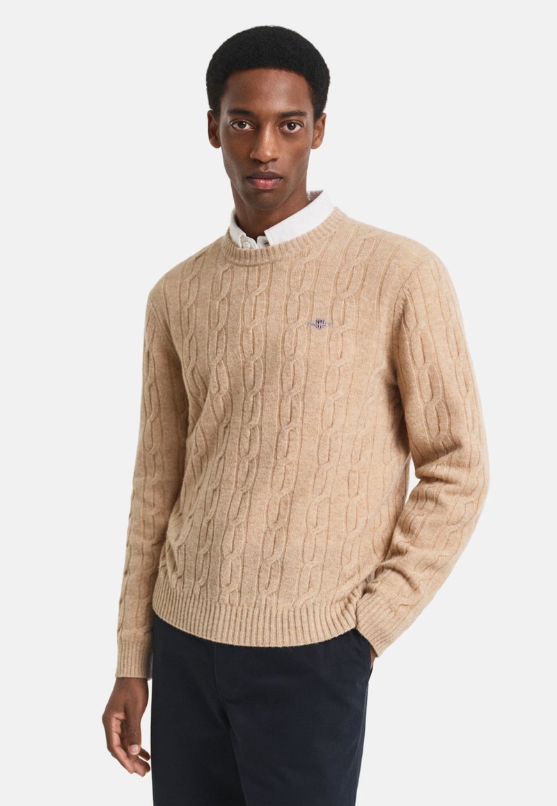 Beige kabelgebreide trui van zacht materiaal met een geribbelde halslijn en zoom, voorzien van een klein logo op de borst. Droeg hij/zij over een wit overhemd met kraag.