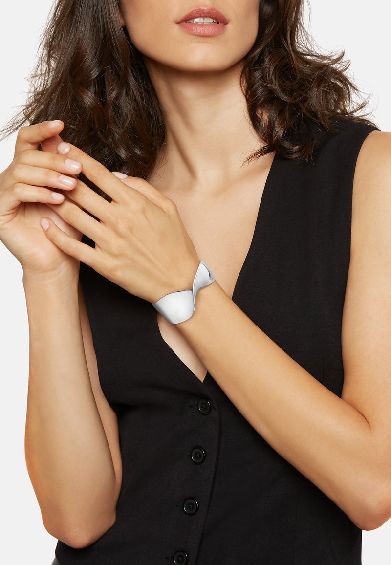 Silverfärgad armband med en slät, vriden design. Materialet verkar metalliskt och reflekterande. Bärs på ett handled, accentuerad av ett svart plagg.
