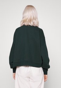 Felpa verde scuro cropped con spalle cadenti, maniche ampie e scollatura rotonda. Tessuto morbido con dettagli di cucitura minimi.