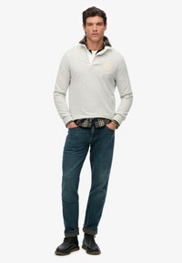 Grå langærmet polo med krave over enternet skjorte, parret med blå jeans og sorte sko, med en afslappet pasform og rullede ærmer.