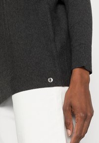 Grauer gerippter Pullover in lässiger Passform, mit einem Saum-Tag, der ein kreisförmiges Logo zeigt, kombiniert mit einem weißen Stoff darunter.