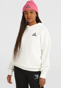 Biely sveter s kapucňou a trojuholníkovým logom, v kombinácii s čiernymi nohavicami s farebnými grafikami. Rôžová štruktúrovaná čiapka dotvára celý outfit.