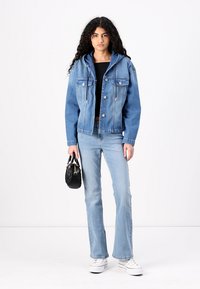 Ανοιχτό μωβ τζάκετ από τζιν με κουκούλα, διαθέτοντας εμπρόσθιες τσέπες και κουμπιά με κλίπ. Φορεμένο πάνω από ένα μαύρο τοπ, συνδυασμένο με ανοιχτά τζιν και λευκά αθλητικά παπούτσια.