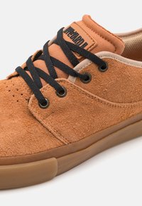 Zapatilla de gamuza marrón con cordones negros, detalles en color tostado, un collar acolchado y una suela de goma. Marca visible en la lengua.