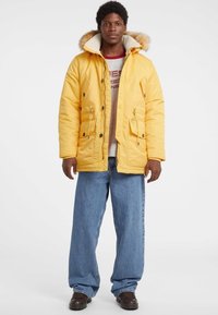 Geel geïsoleerde jas met een nep bontgevoerd capuchon, rits- en drukknoopsluiting, twee voorzakken, gedragen over een lichtgrijs sweatshirt en blauwe jeans.