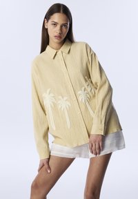 Camisa amarilla texturizada con bordado de palmeras. Manga larga, diseño de botones y corte relajado. Llevada sobre pantalones cortos blancos.
