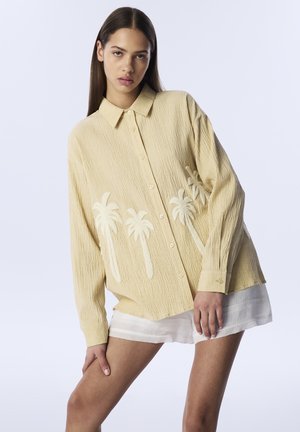 Camicia gialla, con texture, caratterizzata da ricami di palme. Maniche lunghe, design con bottoni e vestibilità comoda. Indossata sopra pantaloni corti bianchi.