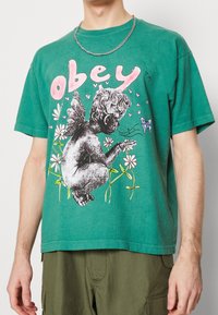 Zelené bavlněné tričko se grafickým potiskem andělské postavy mezi květinami a motýly, s nápisem "obey" v tučných růžových písmenech.