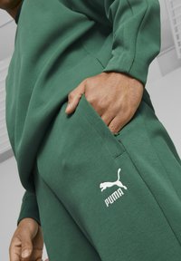 Grüner Sweatshirt und Hose aus strukturiertem Stoff, mit einem weißen Puma-Logo auf der Seitenasche. Lockere Passform mit sichtbaren Nahtdetails.