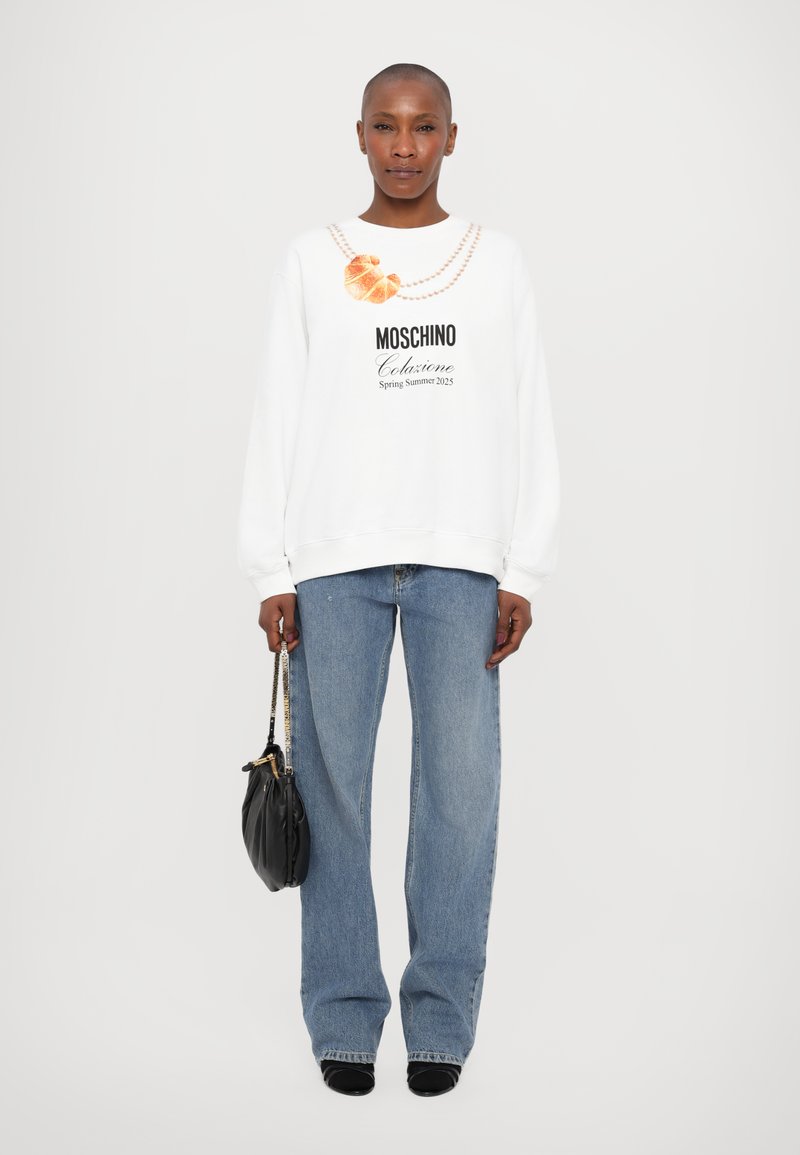 Sweatshirt blanc avec un design graphique représentant une chaîne et un croissant, associé à un jean large bleu clair et un sac à main noir.