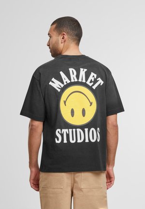 Svart bomull t-skjorte med et stort gult smilefjes-trykk og tekst i hvitt "MARKET STUDIOS" på baksiden. Standard passform med korte ermer.