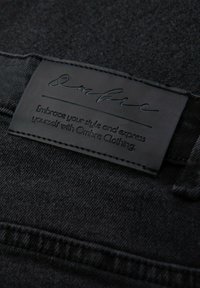 Mustaa denim-kangasta, jonka pinnassa on tekstuuri, sisältää mustan nahkalevyn, johon on painettu brändin nimi "Ombre" ja hoito-ohjeet.