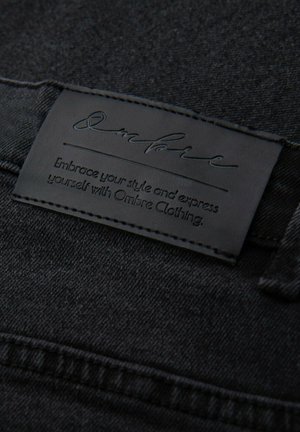 Un tissu en denim noir avec une surface texturée présente une étiquette en cuir noir embossée du nom de la marque "Ombre" et des instructions d'entretien.
