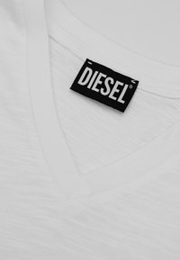 Camicia bianca con scollo a V e un'etichetta rettangolare nera cucita all'interno del colletto che mostra il nome del marchio "DIESEL" in lettere bianche.