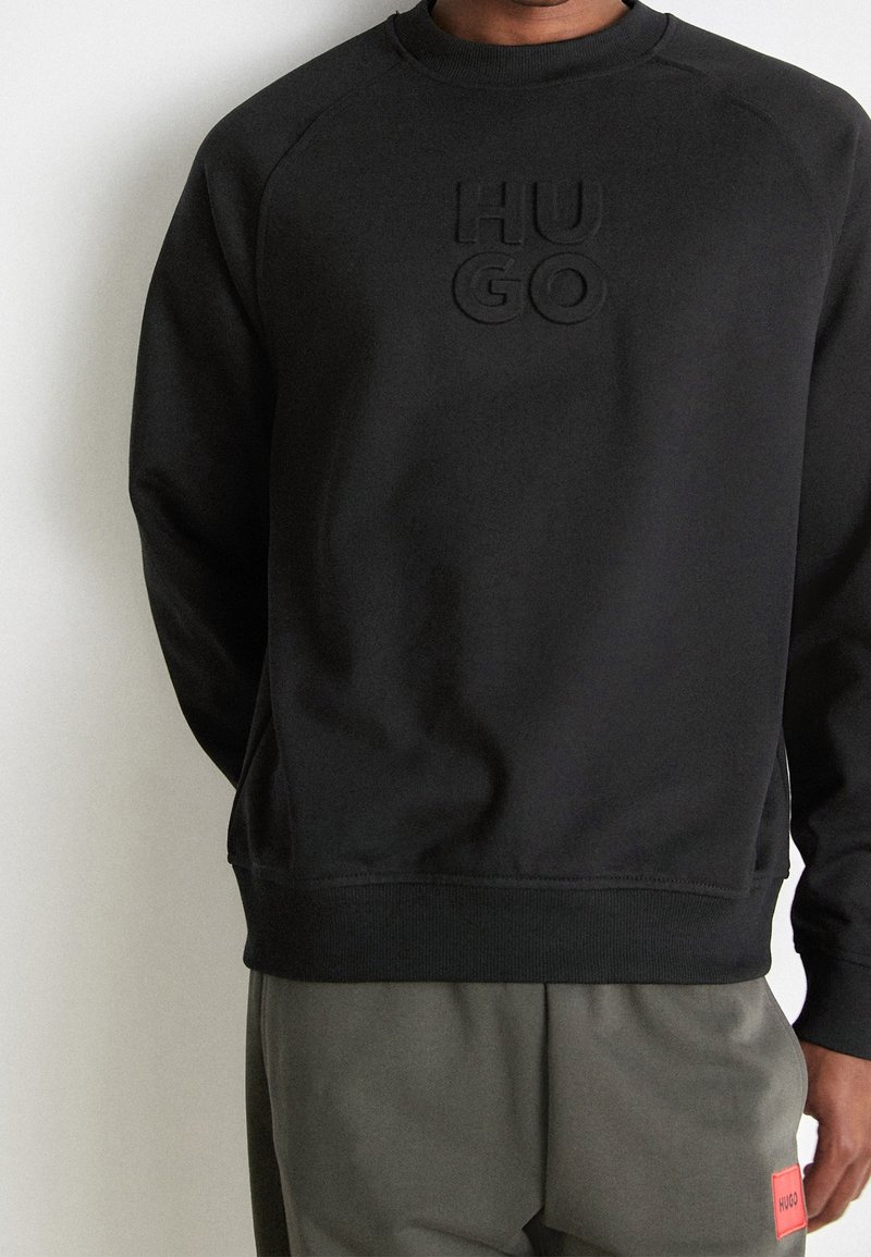 Sweat-shirt noir à manches longues avec logo "HUGO" en relief sur la poitrine, porté par une personne en pantalon gris avec un patch rouge sur la poche.