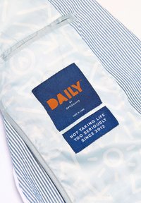 Gaiši zils audums ar baltām svītrām. Satur zilu birku ar uzrakstu "DAILY" oranžā krāsā; zem tā ir teksts: "NAV ŅEMOT DZIVI PĀR TIĶI SERIĀLI KOPŠ 2012. GADA."