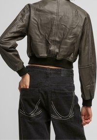 Veste courte en cuir brun avec des poignets et un ourlet côtelés, portée sur un jean en denim foncé présentant des détails de poche en couture blanche.