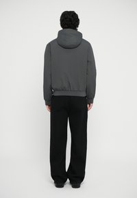 Grå hettegenser med elastisk midje og ermer, kombinert med svarte vide bukser, sett fra ryggen mot en hvit bakgrunn.