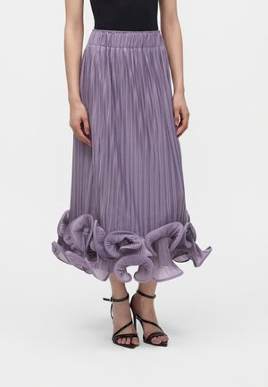 Femme portant une jupe midi plissée lavande avec un ourlet à volants et des sandales à talons hauts noires à lanières.
