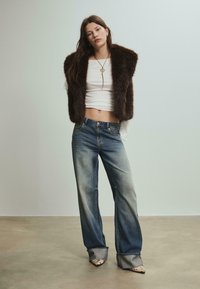 Hnědá umělá kožešinová vesta přes bílý crop top, spárována s vybledlými širokými modrými džíny a špičatými střevíci s leopardím potiskem. Hlazené textury jsou patrné.