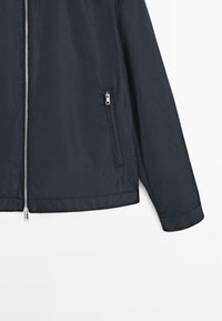 Mørk marineblå jakke med lynlås, lange ærmer, lodret lynlåslomme foran og sølvfarvet lynlås, der løber ned midt foran.