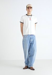 T-shirt blanc avec bordure noire, logo rouge au centre, jeans baggy bleu clair et chaussures à bout pointu marron. Casquette beige et collier inclus.