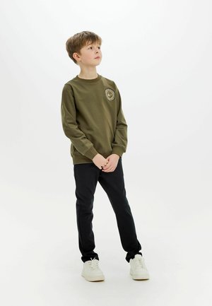 Oliwkowy sweatshirt z okrągłą naszywką na klatce piersiowej, w połączeniu z czarnymi spodniami i białymi sneakersami. Prosty, casualowy design.