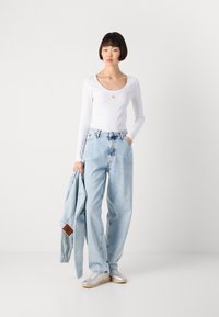Haut blanc à manches longues avec un décolleté arrondi, jeans taille haute bleu clair, et chaussures argentées. Une veste en denim est drapée sur le bras.